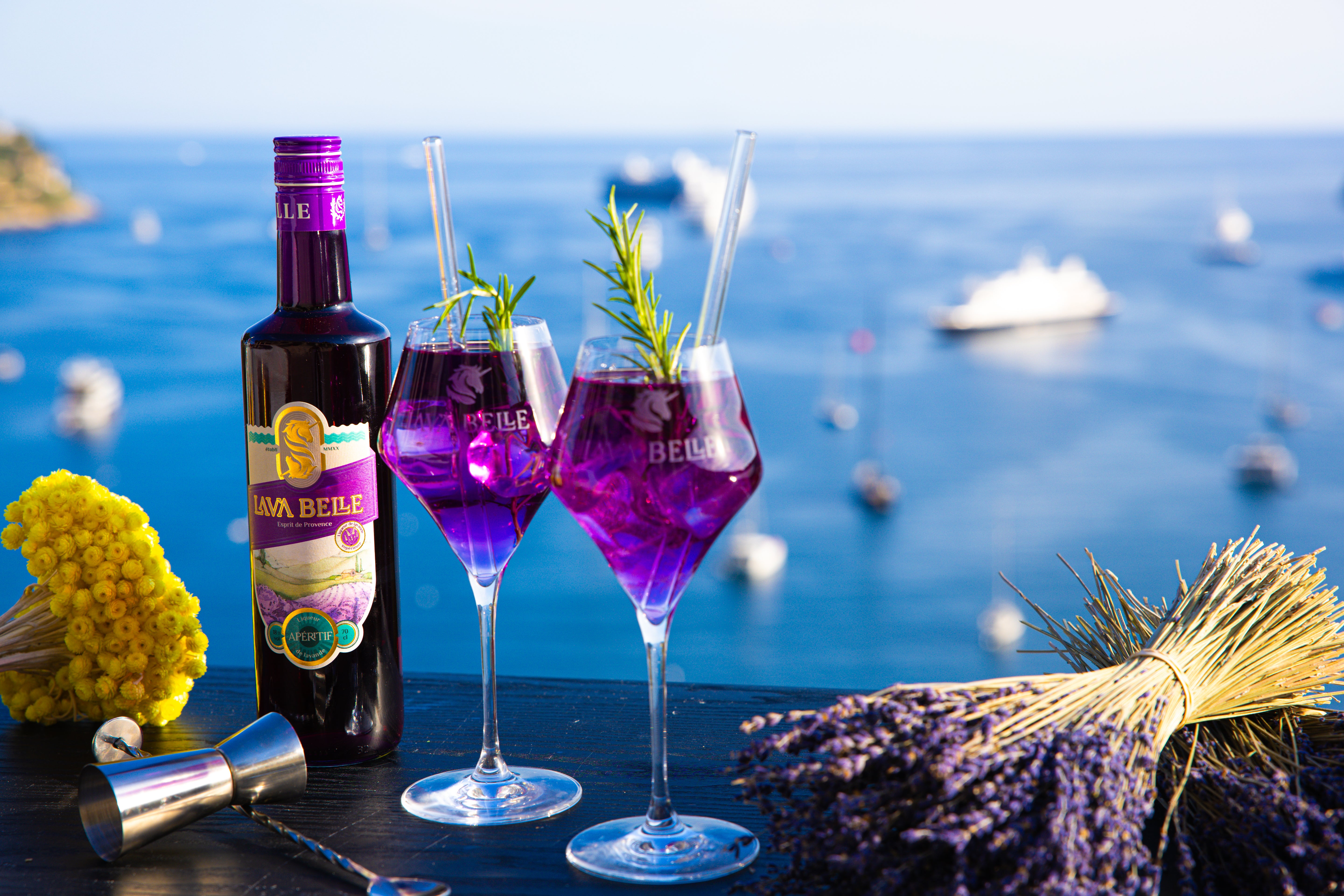 Zwei Gläser Lav'a Belle Spritz sowie eine Flasche vor dem Hintergrund der französischen Riviera mit einigen Botten