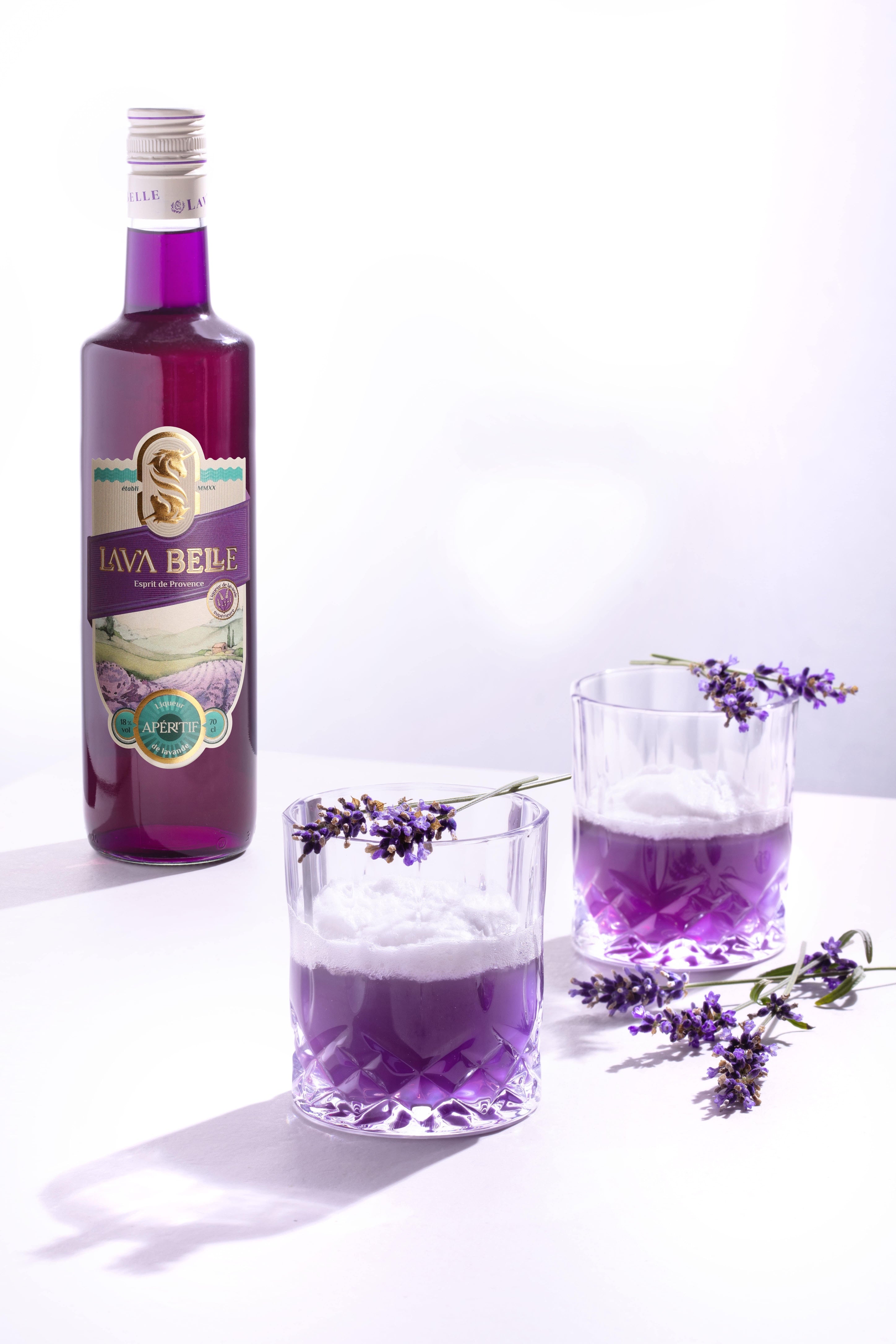 Zwei Tumbler Lav'a Belle Sour mit Schaumkrone und Lavendelzweigen als Dekoration vor einer Flasche Lav'a Belle
